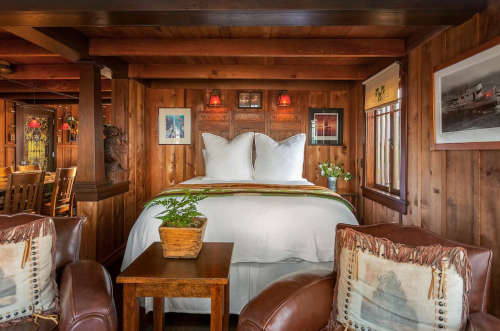 Cozy wooden bedroom with a large bed, two armchairs, and decorative artwork on the walls. Warm, inviting atmosphere.