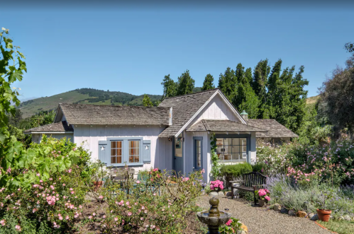 Charming cottage surrounded by colorful flowers and greenery, with mountains in the background under a clear blue sky.