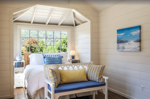 Cozy bedroom with a white bed, blue accents, and a bench by a large window overlooking greenery.