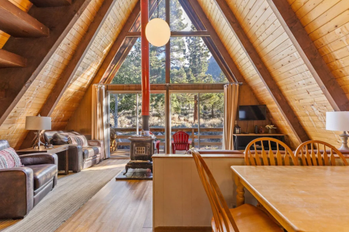 Cozy A-frame cabin interior with wooden beams, a stove, and large windows overlooking nature.
