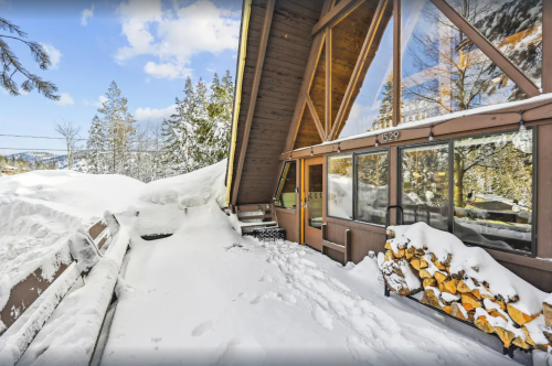 A snowy landscape with a large A-frame cabin and stacked firewood, surrounded by trees under a blue sky.