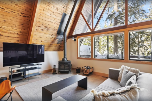Cozy living room with wooden beams, large windows, a TV, and a wood stove, overlooking a snowy landscape.