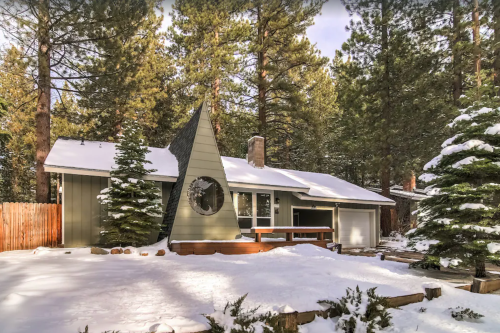 A modern house with a unique triangular roof, surrounded by snow and tall pine trees.