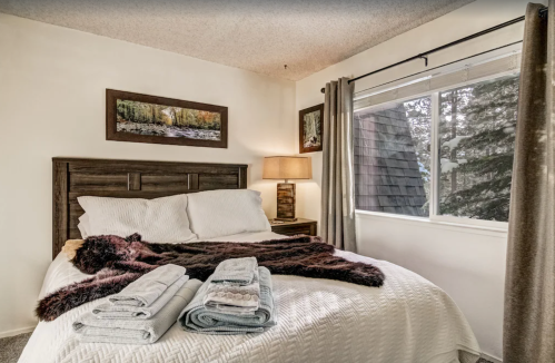 Cozy bedroom with a bed, neatly arranged towels, and a window overlooking trees. Warm lighting adds a welcoming touch.