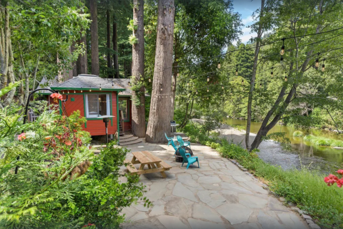Cozy cabin by a river, surrounded by tall trees and a stone patio with outdoor seating and colorful flowers.
