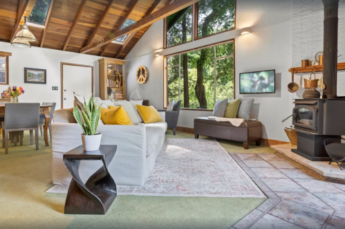 Cozy living room with a sofa, dining area, large windows, and a wood stove, surrounded by greenery.