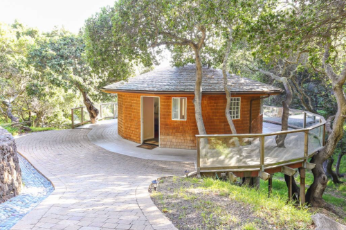 A round wooden cabin surrounded by trees, with a curved pathway leading to its entrance.