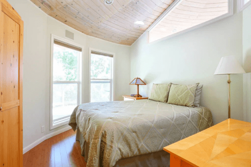 Cozy bedroom with a wooden ceiling, large windows, a bed with decorative pillows, and a lamp on a nightstand.