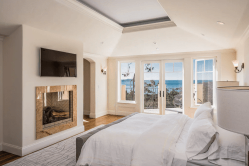 Bright bedroom with a fireplace, TV, and large windows showcasing a scenic ocean view. Elegant decor and natural light.