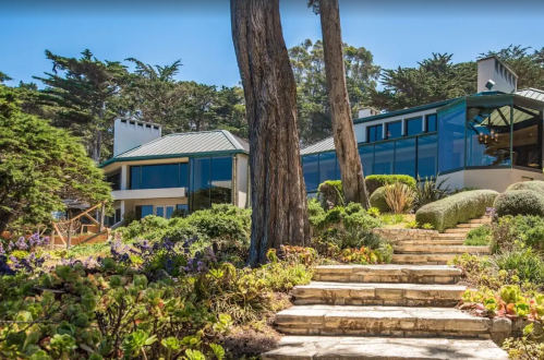 A modern house with large windows, surrounded by lush greenery and stone steps leading to the entrance.