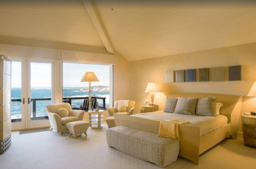 A serene bedroom with a large window overlooking the ocean, featuring a bed, armchairs, and soft lighting.