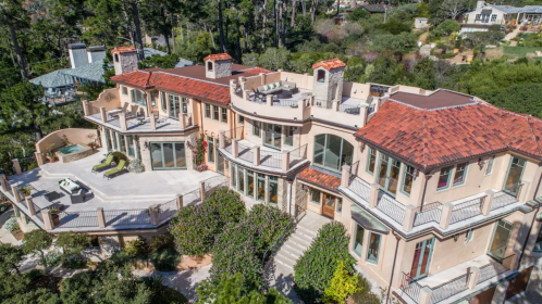 Aerial view of a large, modern home with a red tile roof, surrounded by greenery and outdoor terraces.