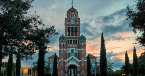 A grand building with a tall tower and domes, surrounded by trees against a colorful sunset sky.