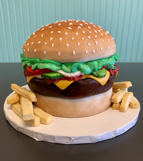 A large, realistic cake shaped like a hamburger, complete with lettuce, tomato, cheese, and fries on the side.