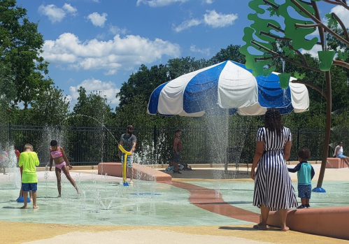 Children play in a splash pad on a sunny day, with water features and a shaded area in the background.
