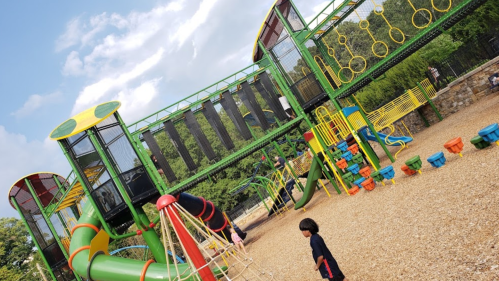 Colorful playground with slides, climbing structures, and swings, surrounded by gravel and green trees.