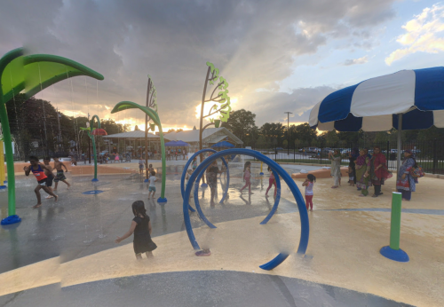 Children play in a splash pad with colorful structures, water features, and a sunset in the background.