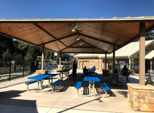 A covered outdoor area with blue picnic tables and a ceiling fan, surrounded by a fence and buildings in the background.