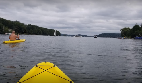 A view from a yellow kayak on a lake, with a person paddling and boats in the distance under a cloudy sky.