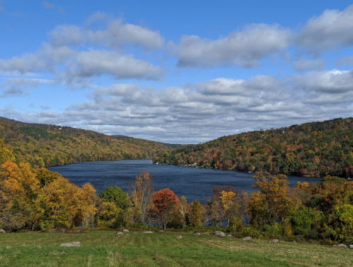 A scenic view of a lake surrounded by autumn foliage and rolling hills under a partly cloudy sky.