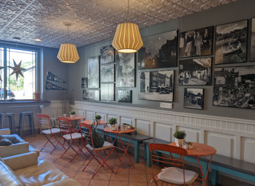 Cozy café interior with orange tables, white walls, and vintage black-and-white photos. Soft lighting from pendant lamps.