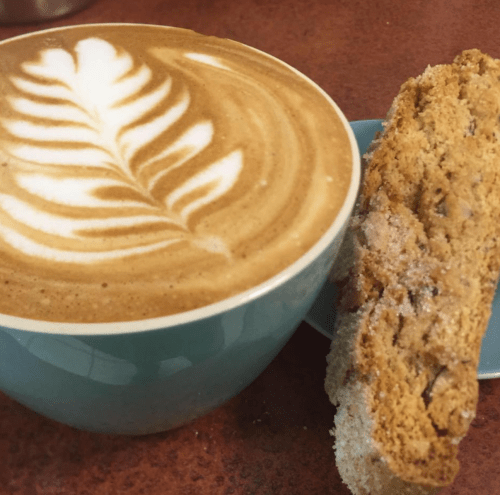 A latte with a leaf design in foam, next to a piece of biscotti on a blue plate.