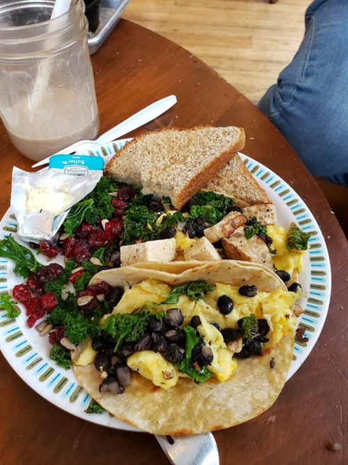 A plate with scrambled eggs, black beans, tofu, kale, dried cranberries, and two slices of whole grain bread.