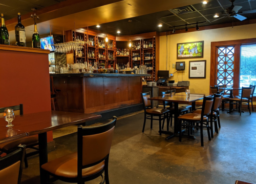 Interior of a restaurant featuring a bar, wooden tables, and shelves lined with bottles. Warm, inviting atmosphere.