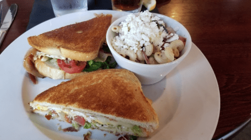 A plate with two toasted sandwiches filled with bacon, lettuce, and tomato, alongside a bowl of pasta salad with feta.