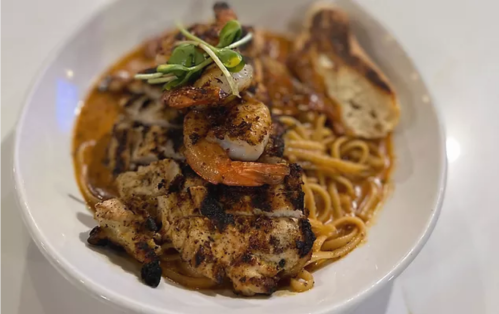 Grilled chicken and shrimp atop pasta in a rich sauce, garnished with greens and served with toasted bread.