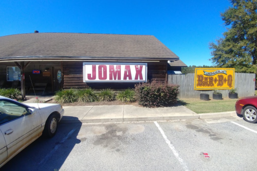 Exterior of Jomax Bar-B-Q restaurant with a wooden facade and signs, parked car in the foreground, clear blue sky.