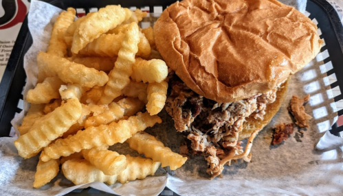 A plate with crinkle-cut fries and a pulled pork sandwich on a bun, served in a black basket with parchment paper.