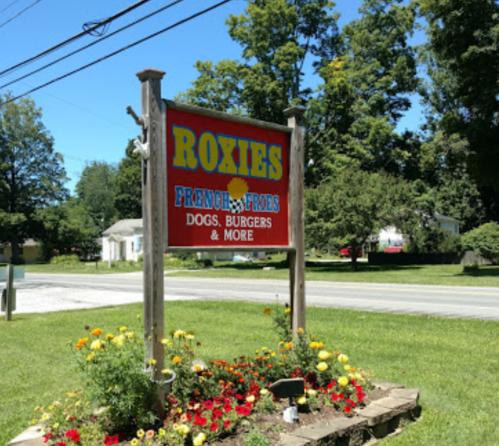 Sign for "Roxie's" featuring French fries, dogs, burgers, and more, surrounded by colorful flowers and greenery.