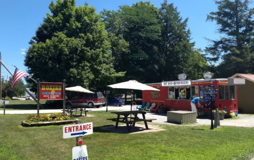 A vibrant food truck with outdoor seating, surrounded by trees and a sunny blue sky, inviting customers to enjoy hot dogs.