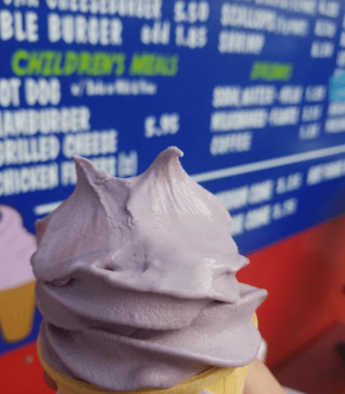 A close-up of a purple soft serve ice cream cone in front of a colorful menu board.