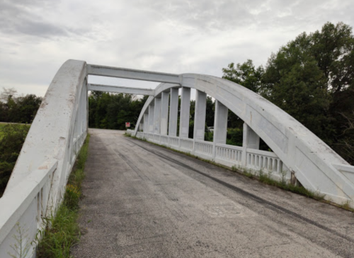 A white arch bridge spans a road, surrounded by greenery under a cloudy sky.