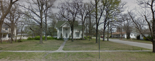 A large white house surrounded by bare trees and a grassy yard, with a quiet street visible in the background.