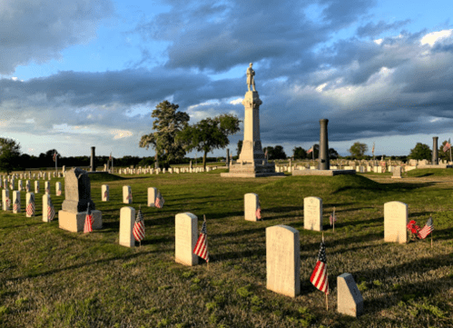 A cemetery with white gravestones, small American flags, and a tall monument under a cloudy sky.