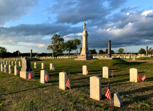 A cemetery with white gravestones, small American flags, and a tall monument under a cloudy sky.