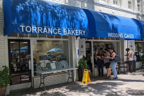 Exterior of Torrance Bakery with blue awnings, people outside, and a table displaying baked goods.