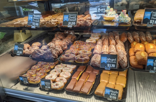 A display case filled with various donuts, including glazed, chocolate, and specialty options, with price tags visible.
