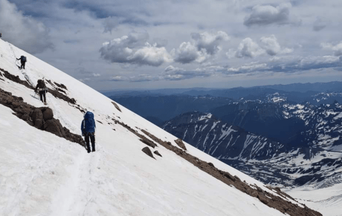 Two climbers navigate a snowy mountain slope under a cloudy sky, with distant peaks visible in the background.