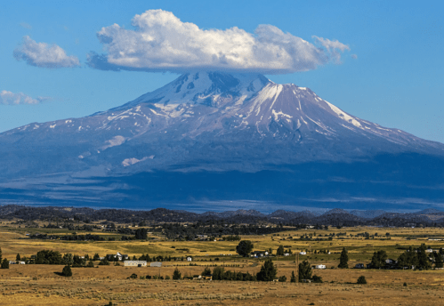 A majestic snow-capped mountain with a cloud cap, surrounded by green fields and distant hills under a clear blue sky.