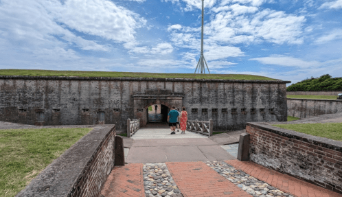 Two people walk towards a historic fort entrance under a partly cloudy sky, surrounded by green grass and stone pathways.