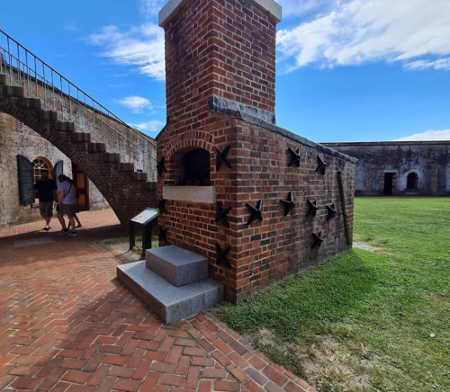 A brick structure with a chimney and metal anchors, near a staircase and grassy area, under a blue sky.