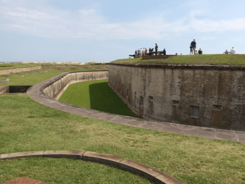 A historic fort with grassy mounds and visitors on top, set against a clear sky.