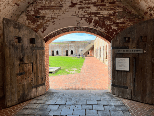 View through an arched doorway of a historic fort, leading to a grassy courtyard with brick pathways.