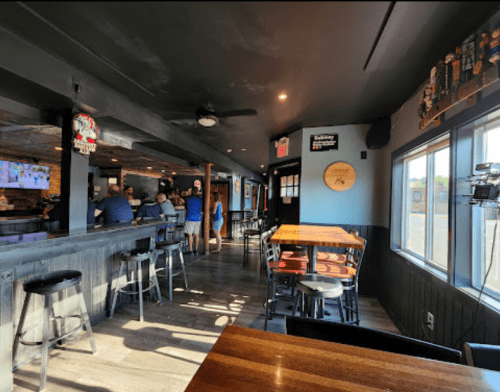 Interior of a bar with wooden tables, stools, and a view of patrons at the counter. Bright and inviting atmosphere.