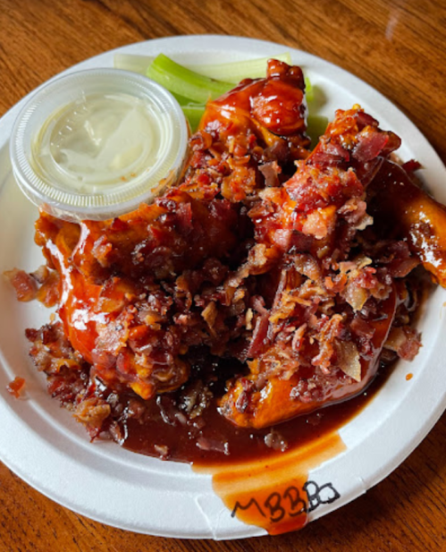 A plate of BBQ chicken wings topped with crispy bacon bits, served with a side of ranch dressing and celery.