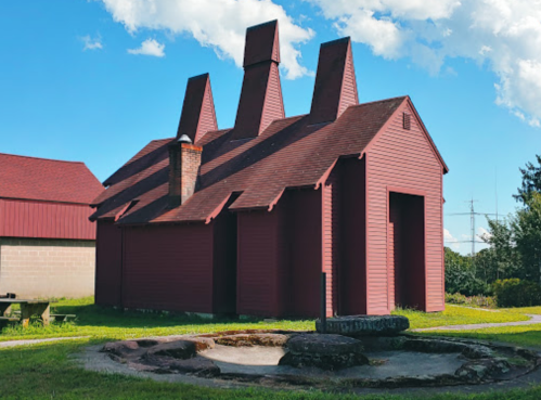 A unique red building with multiple chimneys, set against a blue sky and green grass, with a stone circle in the foreground.